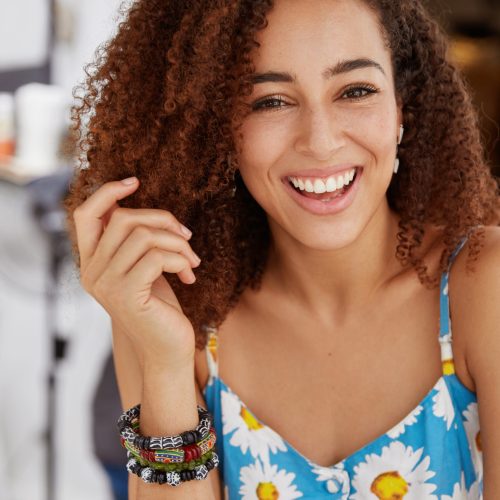 Joyful smiling dark skinned female with bushy hairdo, wears summer t shirt and bracelet, happy to spend free time in family circle or with friends, sits against cafe interior. Happiness concept
