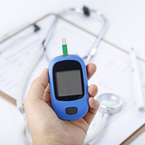 Hand holding a blood glucose meter measuring blood sugar, the background is a stethoscope and chart file