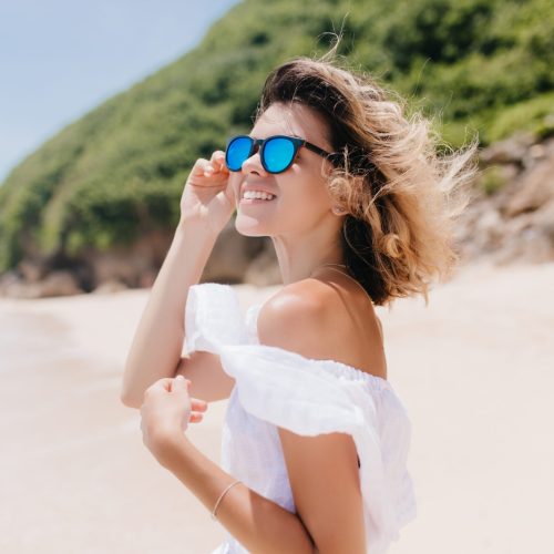 Joyful tanned girl with light-brown hair looking aroud while posing at resort. Magnificent young woman in sunglasses smiling during photoshoot on nature background.
