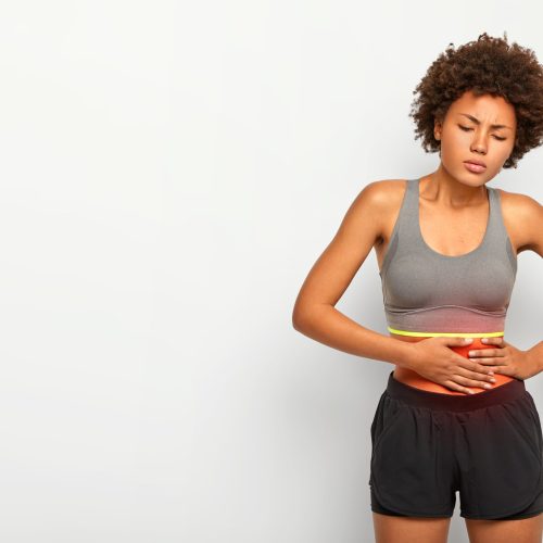 Horizontal shot of dissatisfied African American woman touches belly, suffers from painful cramps, frowns face, dressed in sportclothes, models over white background, free space for your promotion