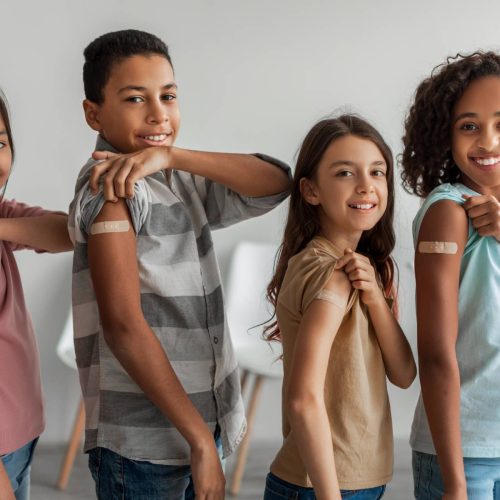 Cheerful Vaccinated Kids Showing Arms After Covid-19 Vaccination Smiling To Camera Posing Indoors. Multicultural Children Group Standing After Receiving Their Vaccine. Coronavirus Immunization